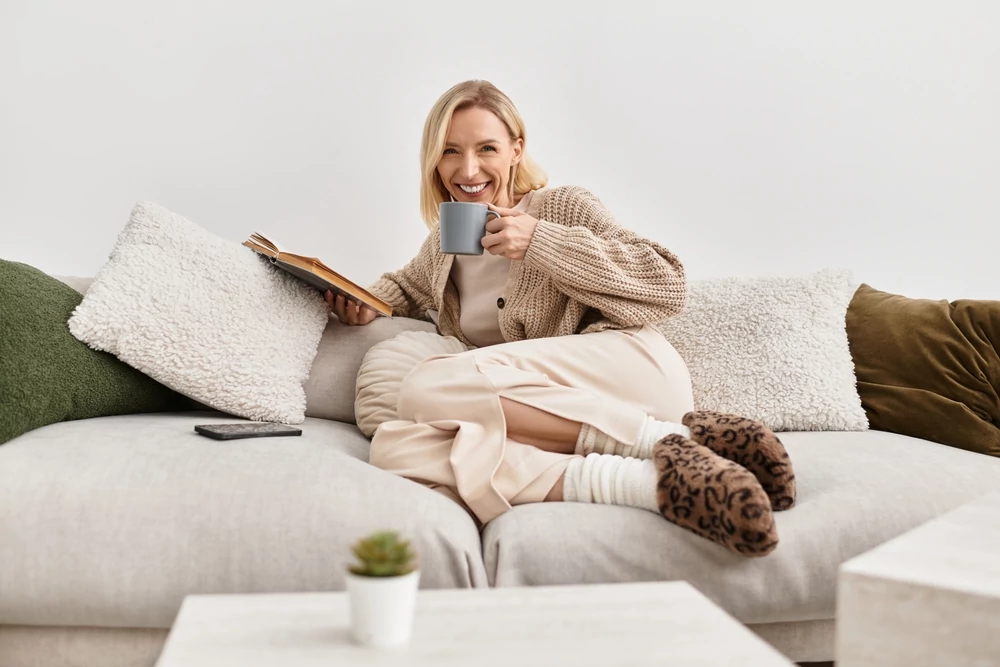 A relaxed blonde woman sips tea while reading a book in a cozy modern living room